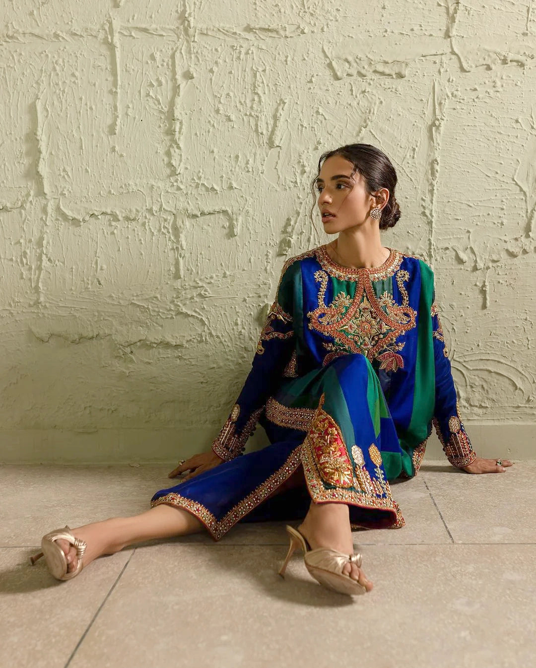 Woman in a blue embroidered traditional outfit sitting against a textured wall.
