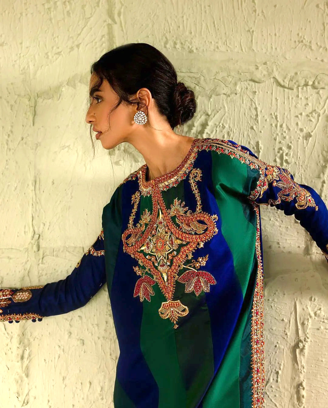 Woman wearing a traditional embroidered outfit against a textured wall.