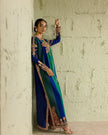 Woman in a blue and green traditional outfit with intricate patterns against a textured wall.