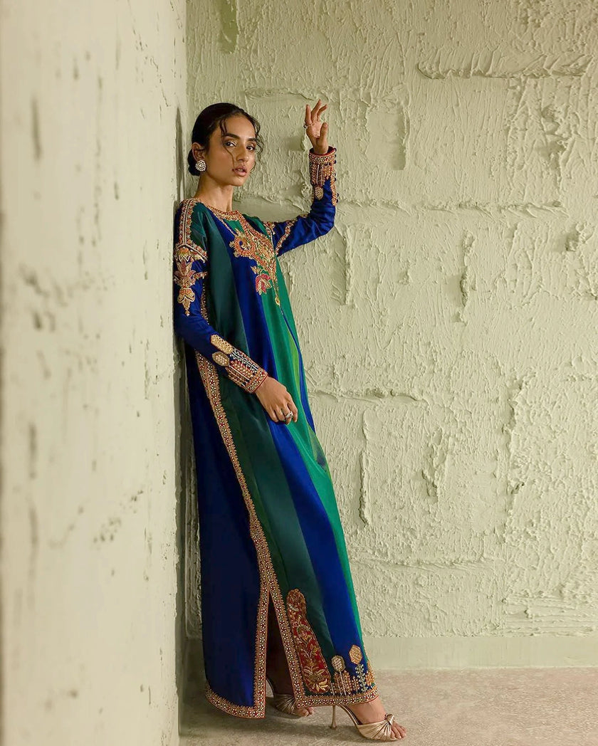 Woman in a blue and green traditional outfit with intricate patterns against a textured wall.