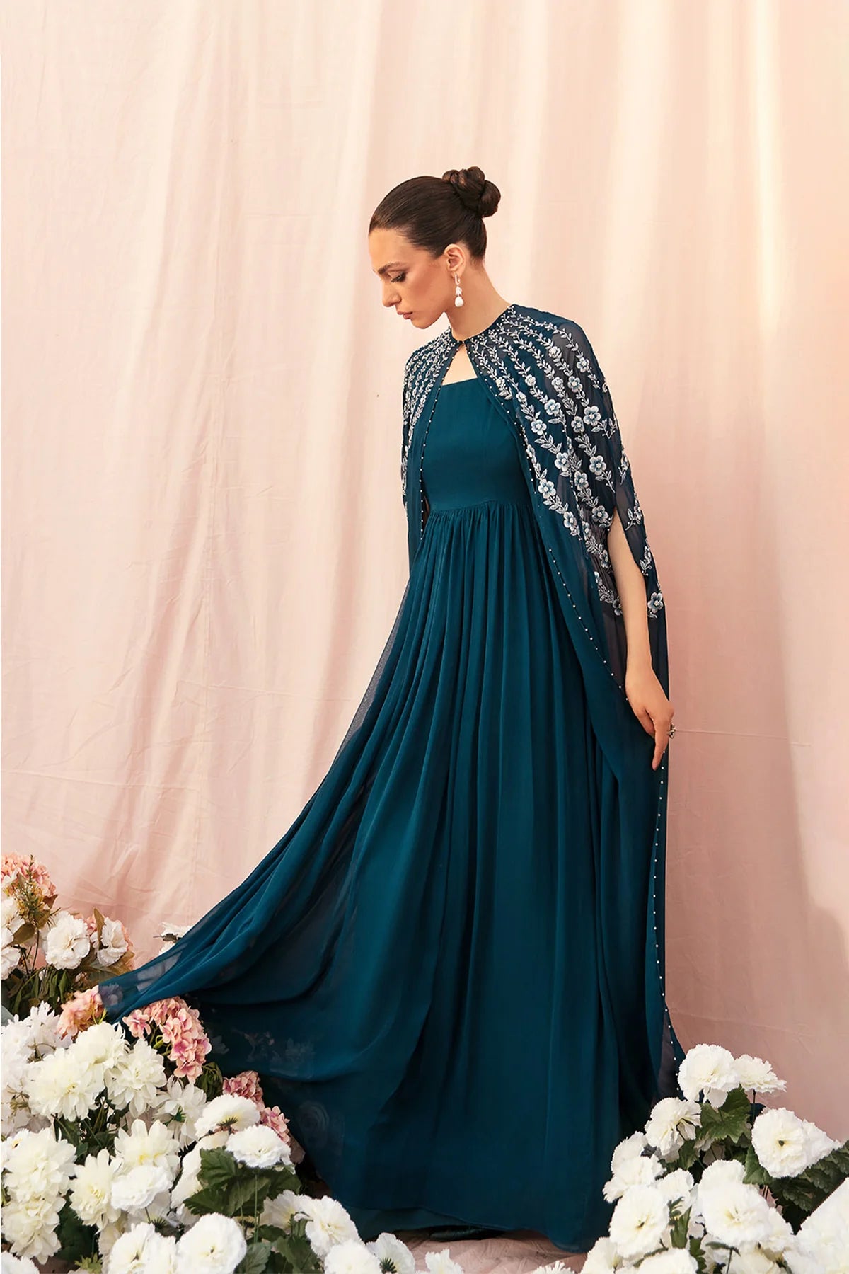 Woman in a teal dress with a matching shawl standing against a pink curtain with floral decorations.