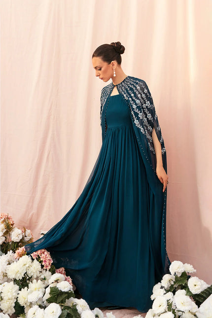 Woman in a teal dress with a matching shawl standing against a pink curtain with floral decorations.